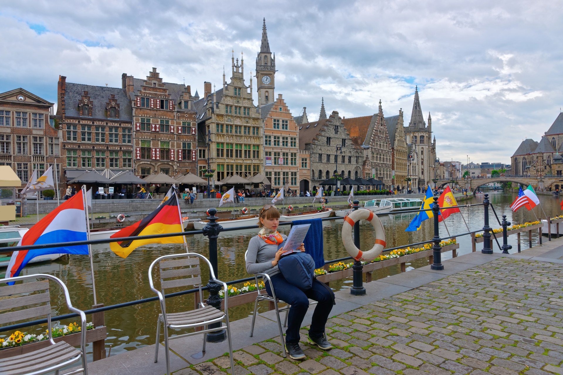 Young woman reading on the Korenlei embankment in Ghent, with the Graslei houses behind her